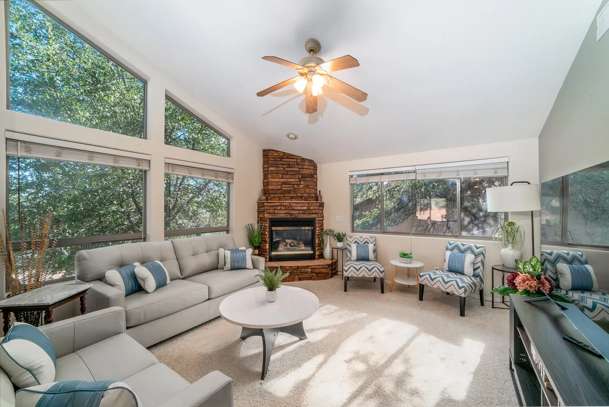 A bright, inviting great room with a stone fireplace, large windows letting in natural light, comfortable seating, and a coffee table. The windows offer views of nature, while the arrangement is perfect for socializing, relaxing, or enjoying a cozy evening indoors.