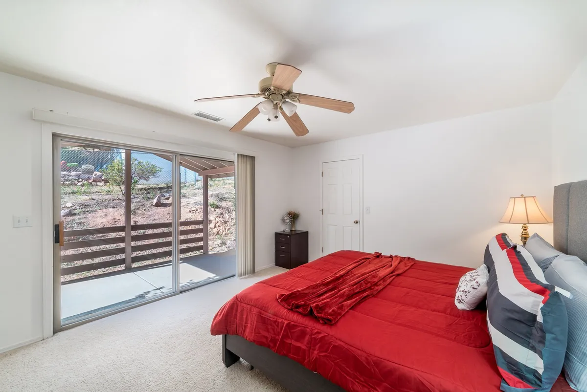 A cozy bedroom with red bedding, a sliding glass door leading to a private patio, a ceiling fan, and a nightstand with a lamp. Natural light fills the room, hinting at serene mornings or starlit evenings just outside.