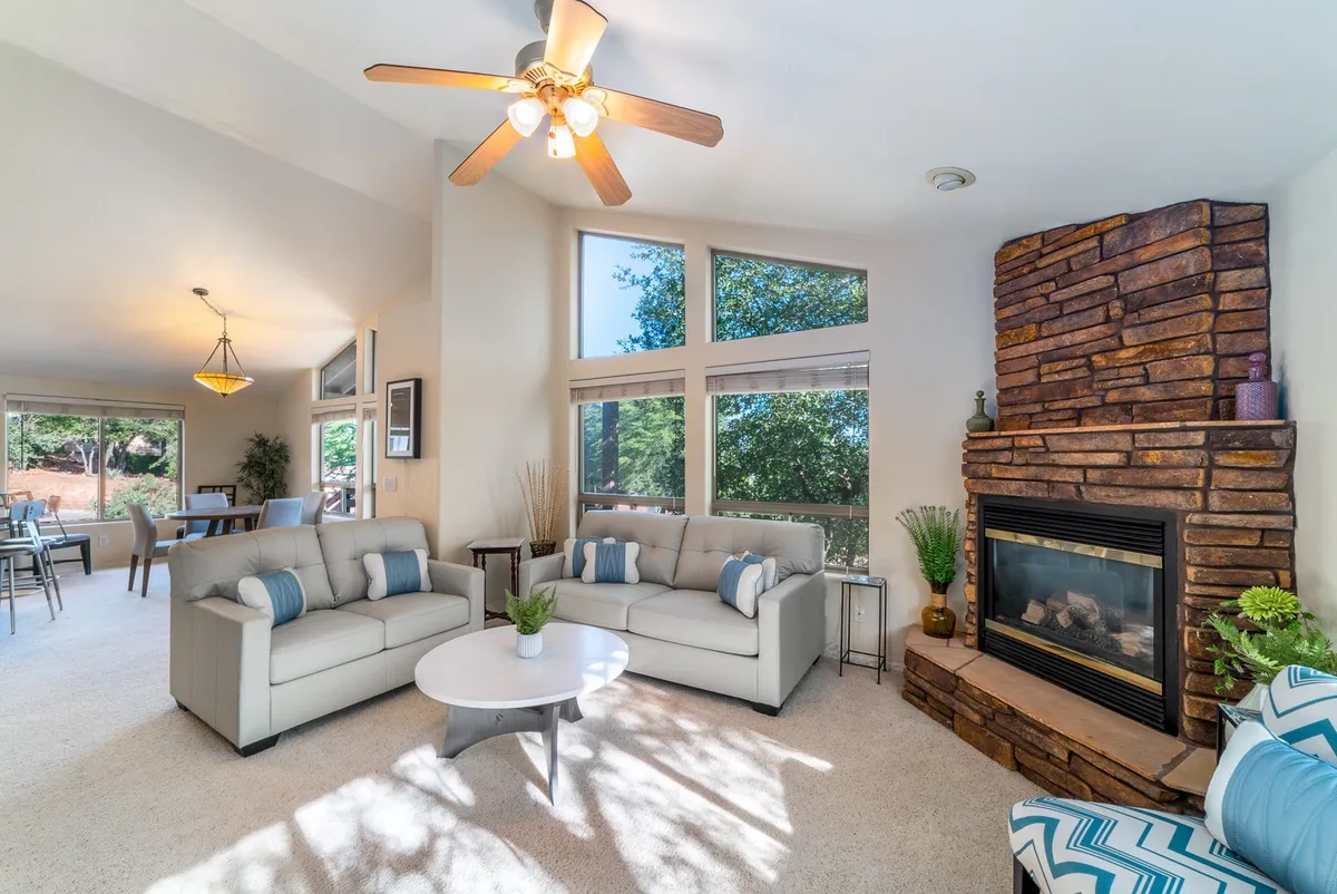 This image captures a spacious living area with comfortable sofas arranged around a fireplace, large windows letting in ample sunlight, and decorative plants enhancing the space’s inviting appeal. The open concept and clear walkways emphasize ease of movement and a welcoming atmosphere for gathering and relaxation.