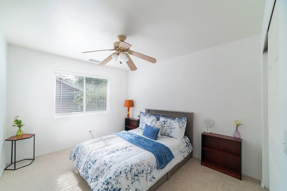 A tranquil bedroom featuring a bed with floral bedding, a ceiling fan, a window letting in soft natural light, decorative side table with flowers, and wooden nightstands. The space feels calm and comfortable—perfect for restful sleep.