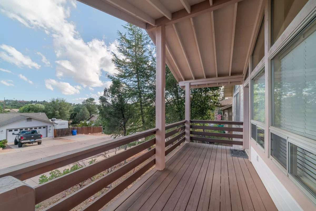 A spacious wooden deck bordered by a sturdy railing, surrounded by lush green trees and open sky. The deck overlooks a sloped landscape with a driveway below. The ambiance suggests a peaceful, elevated vantage point ideal for relaxation and social gatherings. The setup hints at activities like enjoying sunrise coffee, sunset drinks, or hosting a family barbecue in the fresh mountain air.