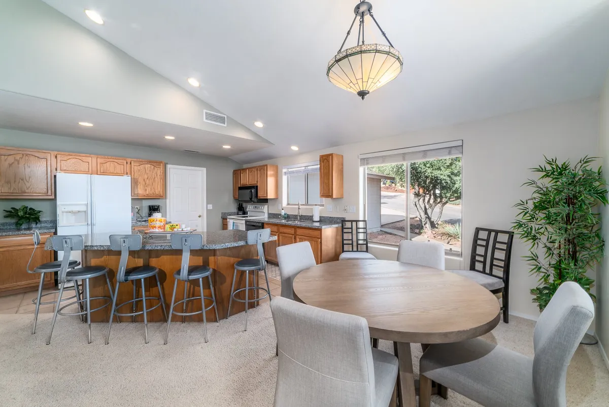 An inviting dining area adjacent to the kitchen, featuring a dining table set for four and a kitchen island in the background. The space is brightened by windows and designed for both intimate meals and festive gatherings.