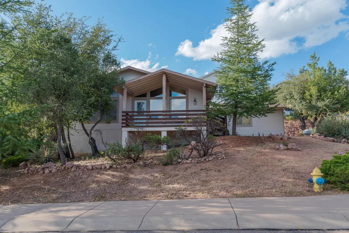 The image features a wooden deck at the main entrance of the house, with a tall pine tree and rich greenery encircling the area. The deck offers an inviting space for outdoor relaxation, complemented by the tranquil setting and ample room for seating or a grill station. The scene evokes a sense of hosting barbecues, enjoying sunsets, or unwinding after outdoor activities.
